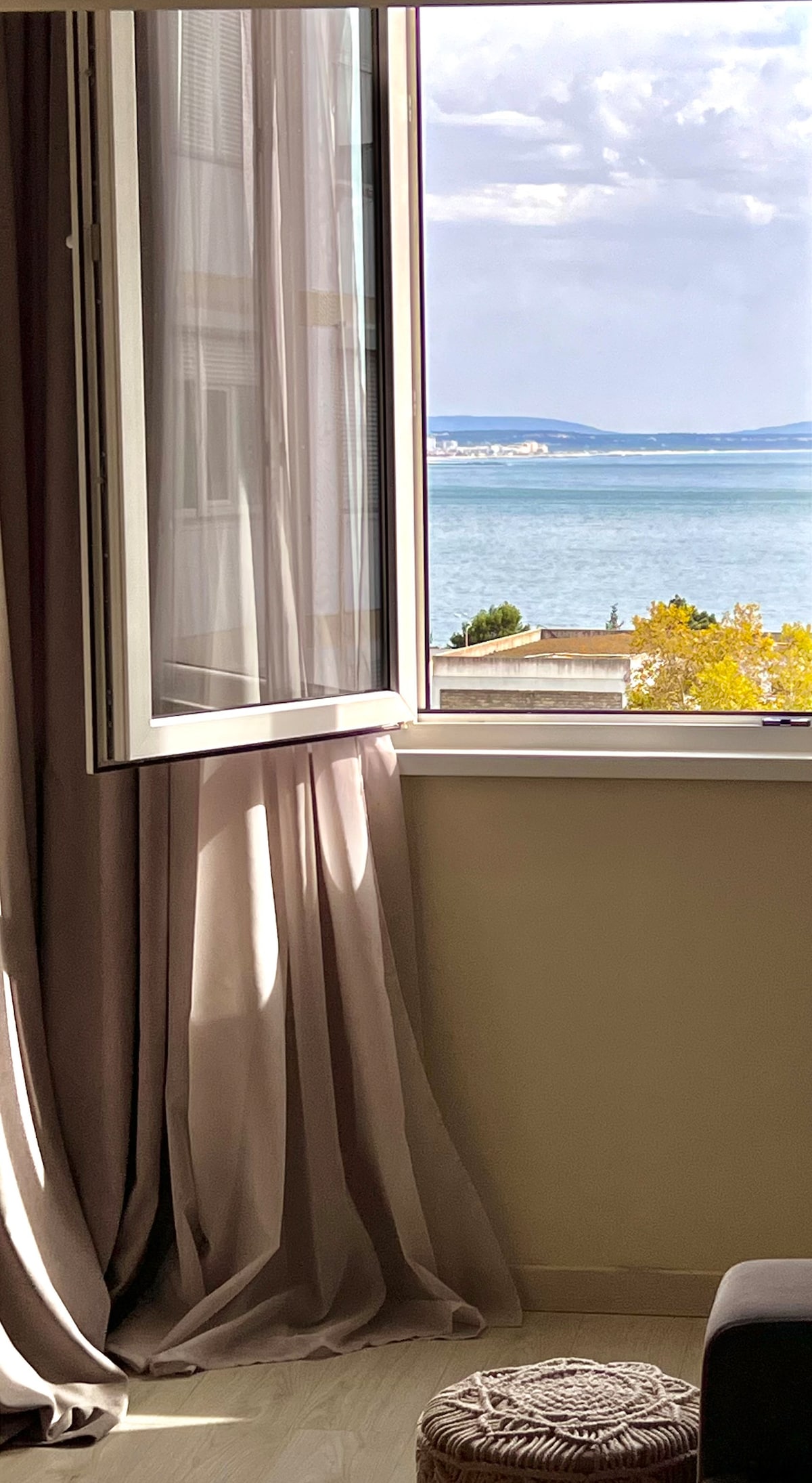 A large window opens to a view of the ocean, with soft, flowing curtains framing the scene. The sunlight illuminates the room, casting gentle shadows on the interior walls. In the foreground, a small decorative pouf is visible on the floor.