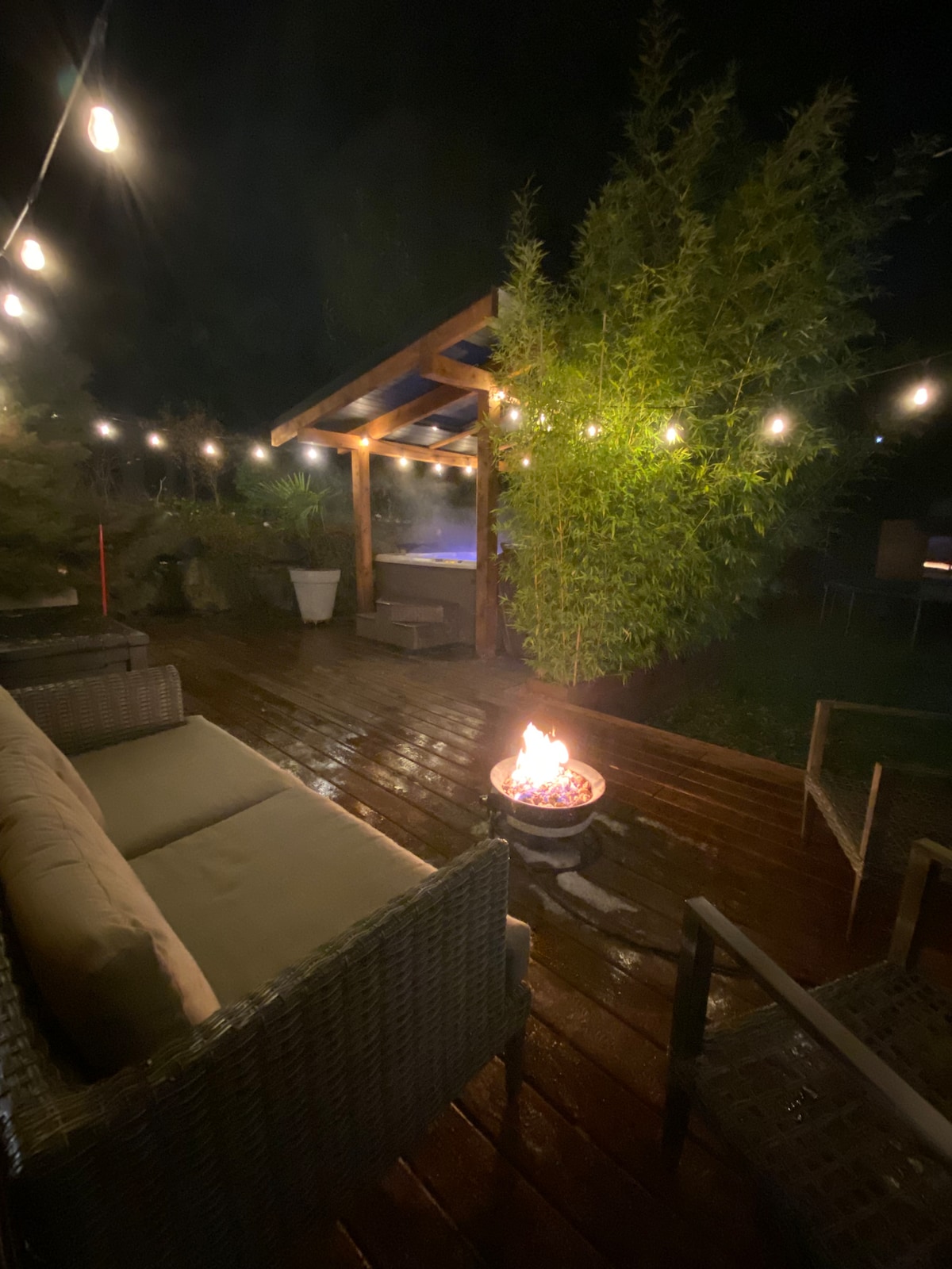 The serene patio area is illuminated by string lights, highlighting a cozy seating arrangement next to a gentle flame in a fire pit. A covered space is visible in the background, providing shelter and enhancing the inviting atmosphere of this outdoor retreat.