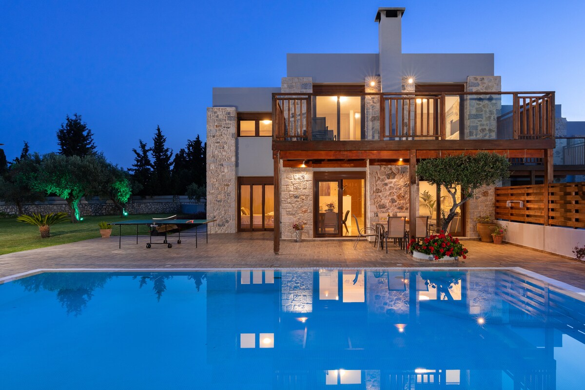 A modern villa is positioned beside a large swimming pool, reflecting the evening sky. Natural stone exterior harmonizes with wooden balconies. Large glass doors illuminate welcoming interior spaces. Lush greenery and blooming flowers enhance the outdoor area, creating a serene atmosphere for relaxation.