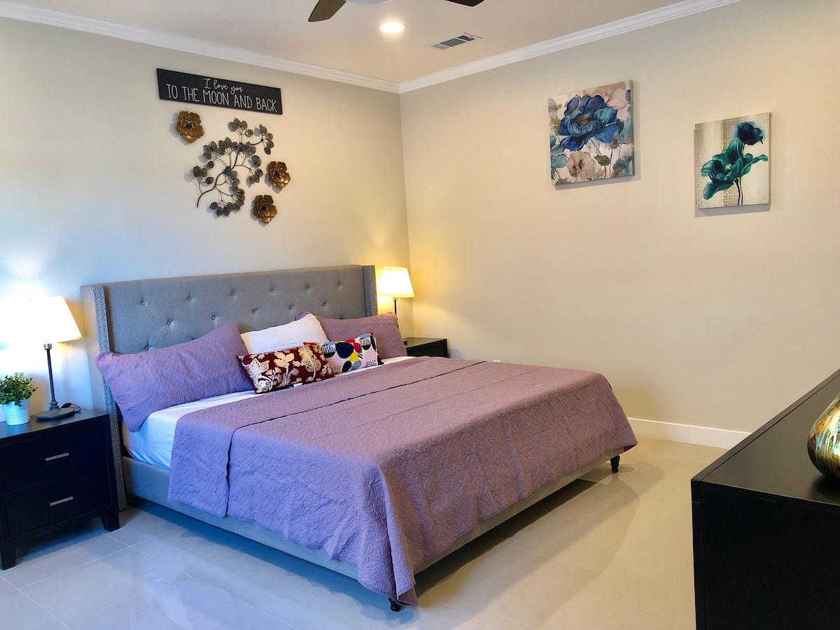 A cozy sleeping area is furnished with a spacious bed covered by a purple blanket. Nightstands with lamps are placed on either side, and wall art featuring floral designs adds a touch of color. Soft lighting creates a soothing atmosphere in the well-appointed room.