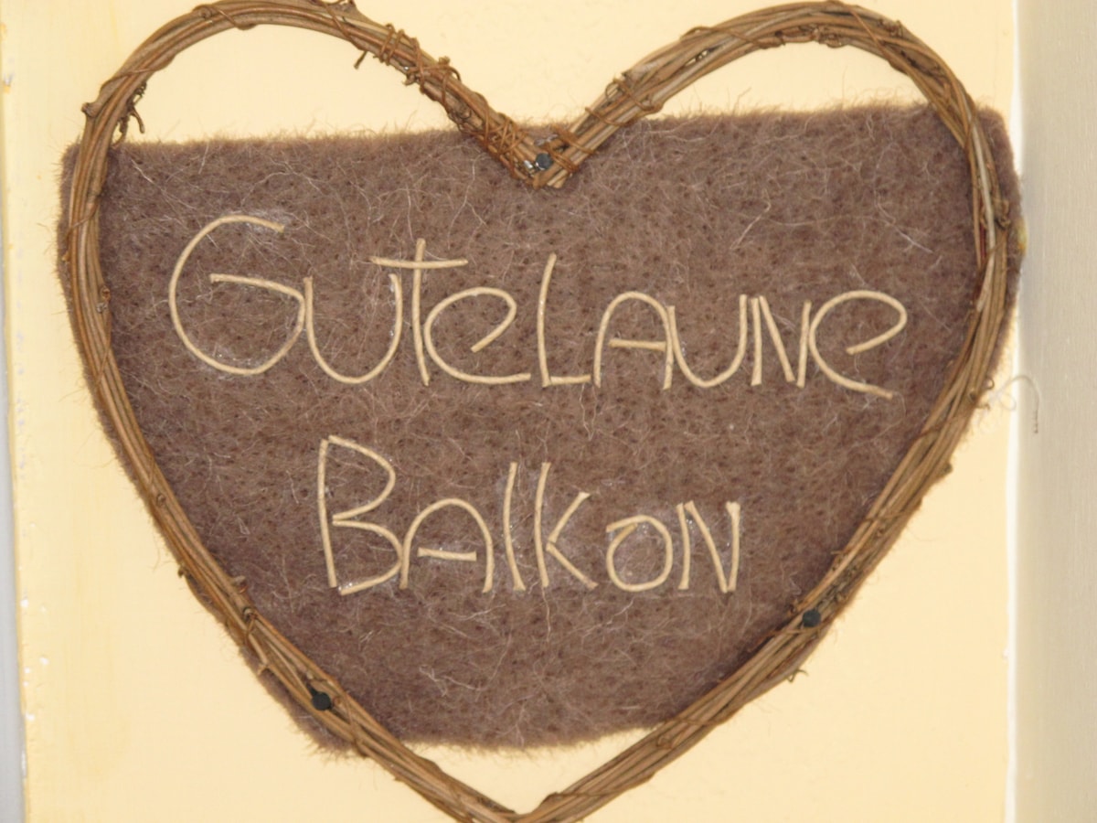 A heart-shaped wall decoration is displayed, featuring the words 'Guten Laune Balkon' crafted from natural materials. The background is composed of a soft, brown fabric, creating a rustic and inviting ambiance.