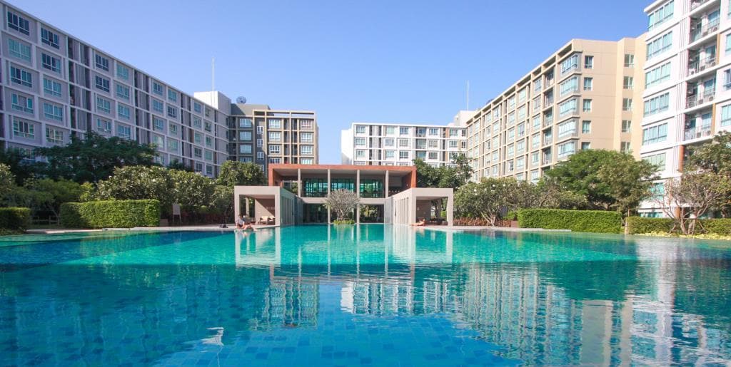 A spacious swimming pool is surrounded by landscaped gardens and modern condominium buildings. The pool's clear blue water reflects the sunny sky, and lounge areas can be seen along the pool's edge, creating a serene outdoor atmosphere.