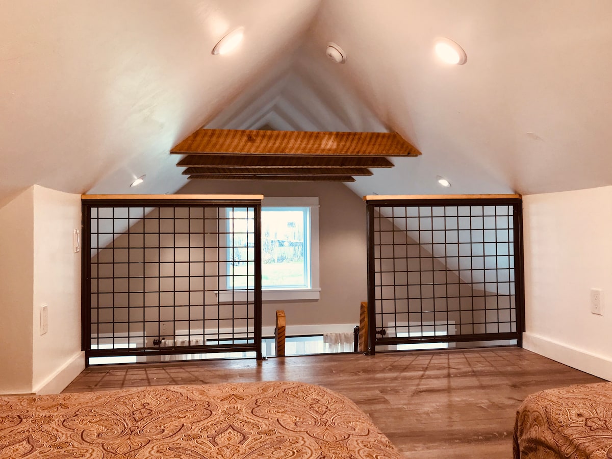 An elevated loft space is showcased, featuring wooden beams along the sloped ceiling. Safety railings with a grid design provide a secure boundary. Natural light filters through a window, illuminating the rich texture of the patterned bedding below.