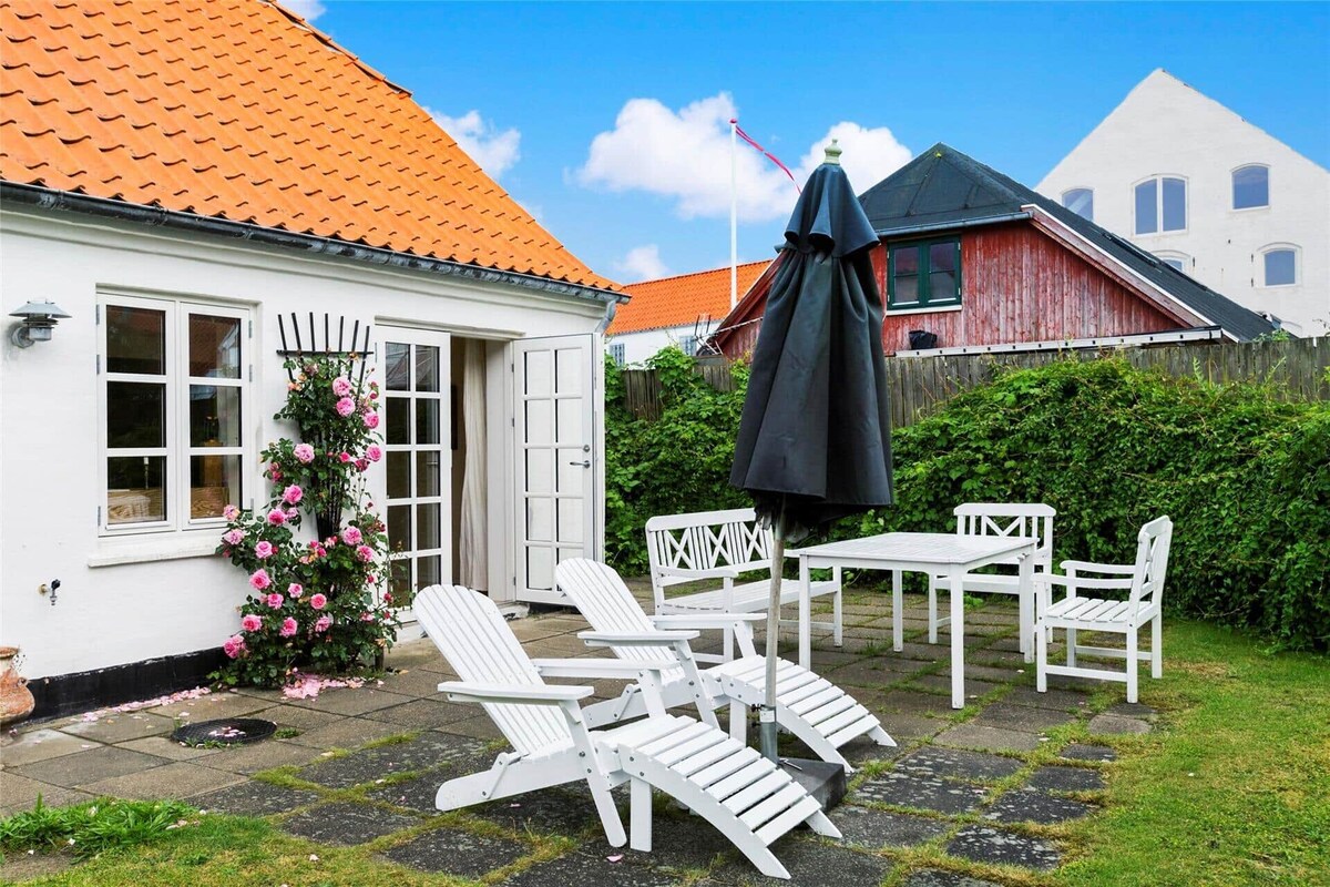 A private garden area features a stone patio adorned with white outdoor furniture, including chairs and a table. A large umbrella provides shade, while climbing roses add a natural touch against the house's white exterior. The bright blue sky complements the inviting space.