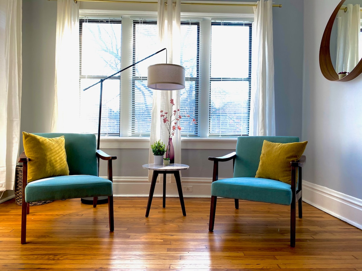 A comfortable sitting area is presented with two teal chairs, each adorned with a yellow accent pillow. A small round table is positioned between the chairs, featuring a decorative plant. Natural light filters through large windows, surrounded by sheer white curtains.