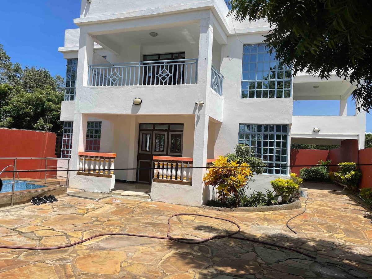 A spacious house exterior is depicted, featuring large windows that allow natural light. The surrounding garden includes colorful plants and a stone pathway. A swimming pool is visible to one side, contributing to the enjoyable outdoor space ideal for relaxation.