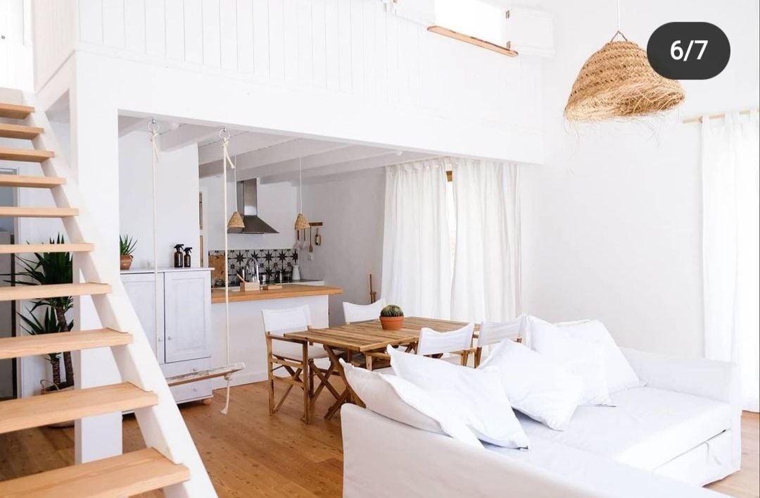 A bright, airy living space features a comfortable white sofa adorned with plush cushions. A wooden dining table with chairs is centrally positioned, while a modern kitchen area is visible in the background, accented by wooden elements. Soft light filters through sheer curtains, enhancing the welcoming environment.