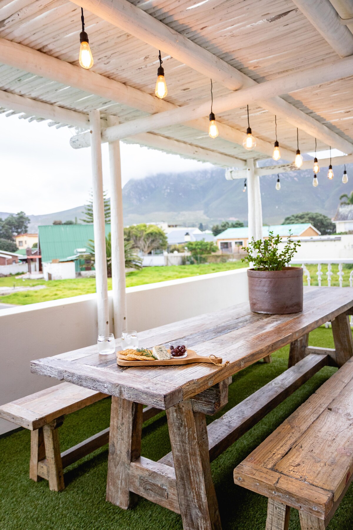 An outdoor dining area is featured, equipped with a rustic wooden table and benches. A potted plant sits at the center of the table, which is adorned with a cheese platter and grapes. String lights hang above, providing subtle illumination.