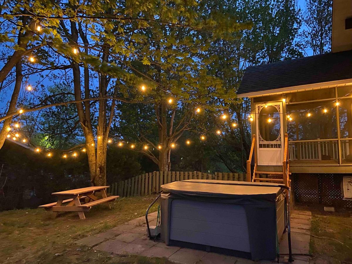 A serene outdoor space is illuminated by string lights in the evening. A hot tub sits on a paved area, surrounded by a grassy yard. A picnic table is positioned nearby, with trees providing a natural backdrop.