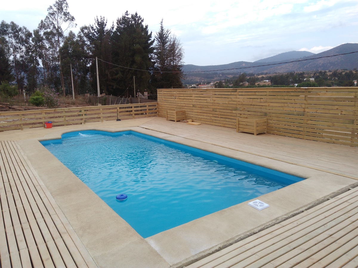 A rectangular pool is surrounded by a wooden deck, offering space for relaxation. The area is enclosed by wooden fencing, with distant views of rolling hills and trees visible in the background. The serene water reflects the sky above.