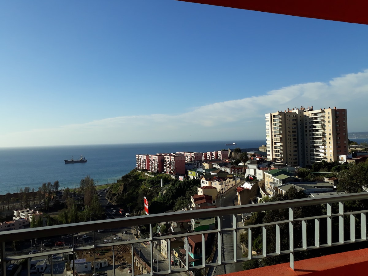 A panoramic view is presented from the terrace, showcasing the coastline and the ocean. Ships are seen in the distance, while urban and natural landscapes blend seamlessly below. The sky is clear with minimal clouds, enhancing the expansive vista.