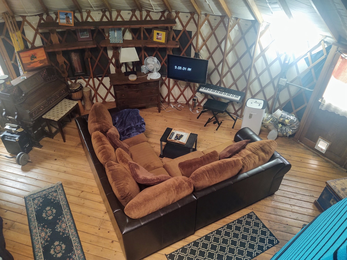 A spacious living area within a yurt features a large sectional couch, facing a low coffee table. A flat-screen TV is positioned alongside a keyboard, while a wooden cabinet and a vintage piano add character to the space. Natural light floods the interior through windows and skylights.