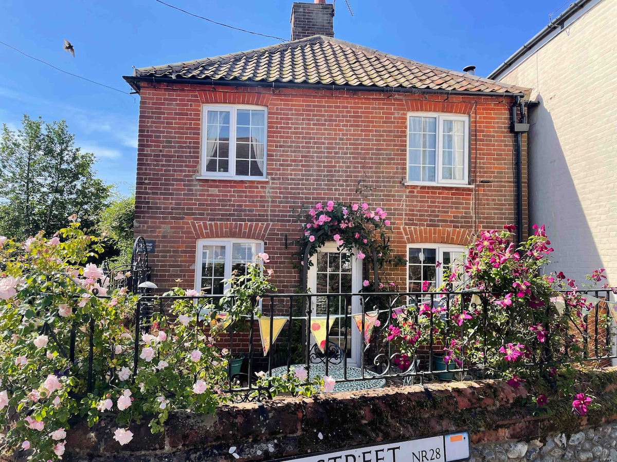 A charming brick cottage features a sweet facade adorned with colorful climbing roses. Large windows allow natural light to fill the interior, while a tasteful wrought-iron fence creates a welcoming entry. The peaceful surrounding garden enhances the tranquil atmosphere of the village street.