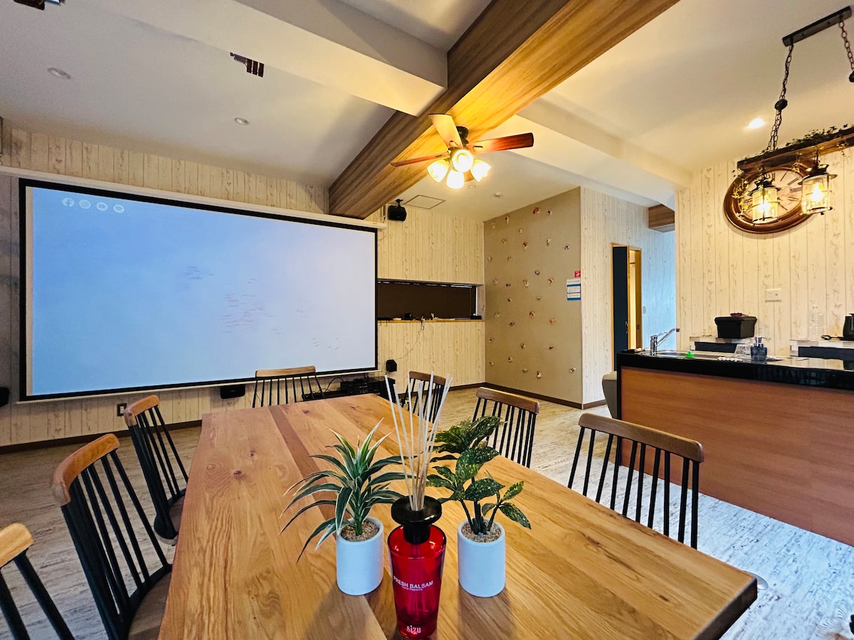 The spacious living area includes a large dining table surrounded by black chairs, complemented by a decorative plant arrangement. A large projector screen is mounted on the wall, while an open kitchen area is visible in the background, enhancing a communal atmosphere.