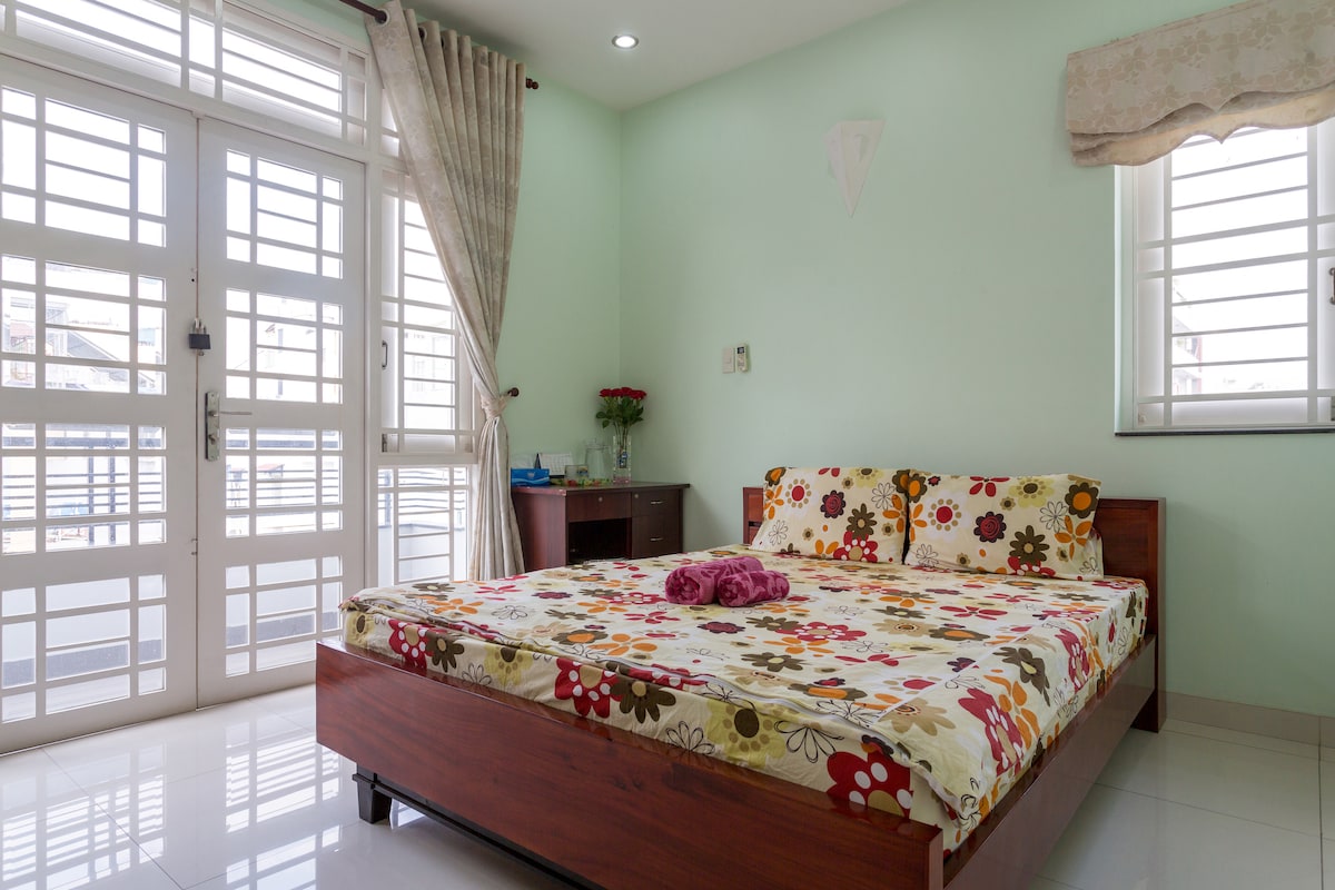 A bright room features a spacious bed adorned with a floral-patterned cover. Natural light filters through large windows, and tasteful curtains frame the scene. A wooden nightstand is positioned beside the bed, enhancing the clean and simple aesthetic of the space.