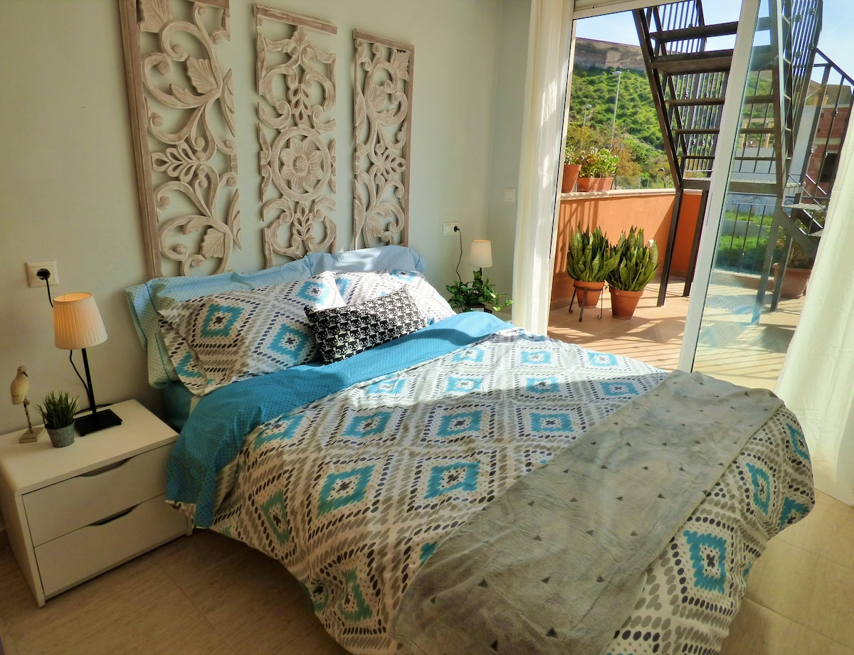 A sleeping area features a spacious bed dressed in a colorful patterned duvet. An intricately carved wooden headboard adds texture, while natural light floods the room through glass doors that open to a terrace. Potted plants can be seen outside, enhancing the serene ambiance.