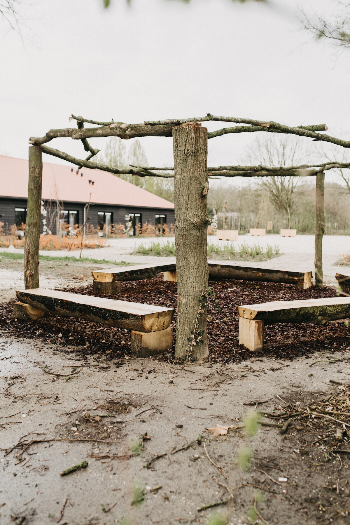 An outdoor seating area features wooden benches arranged in a circular shape around a central tree. Mulch covers the ground beneath the benches, creating a natural gathering space. The surrounding landscape includes trees and a building visible in the background.