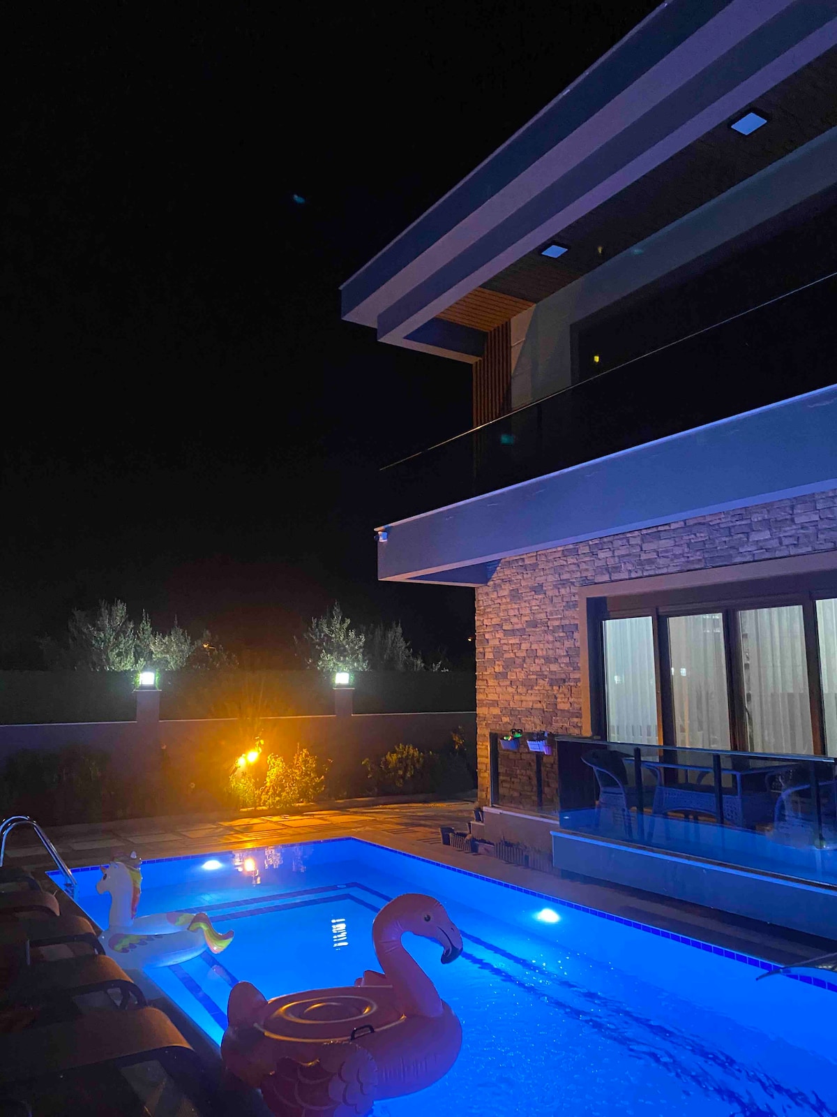 A modern villa stands illuminated at night, showcasing a private swimming pool with soft blue lighting. A floating pink flamingo adds a playful touch to the serene outdoor space, complemented by surrounding greenery and ambient lighting from the garden.