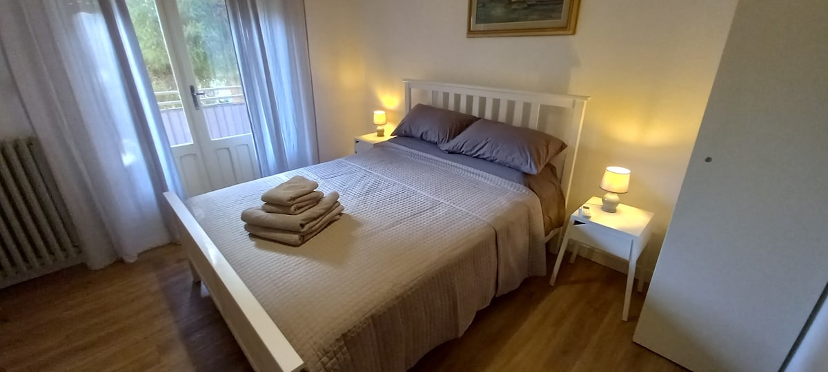 A spacious bedroom features a white bedframe with a neatly arranged quilt and stacked towels. Soft lighting is provided by two bedside lamps, creating a warm ambiance. Large windows with light curtains allow natural light to enhance the inviting setting.