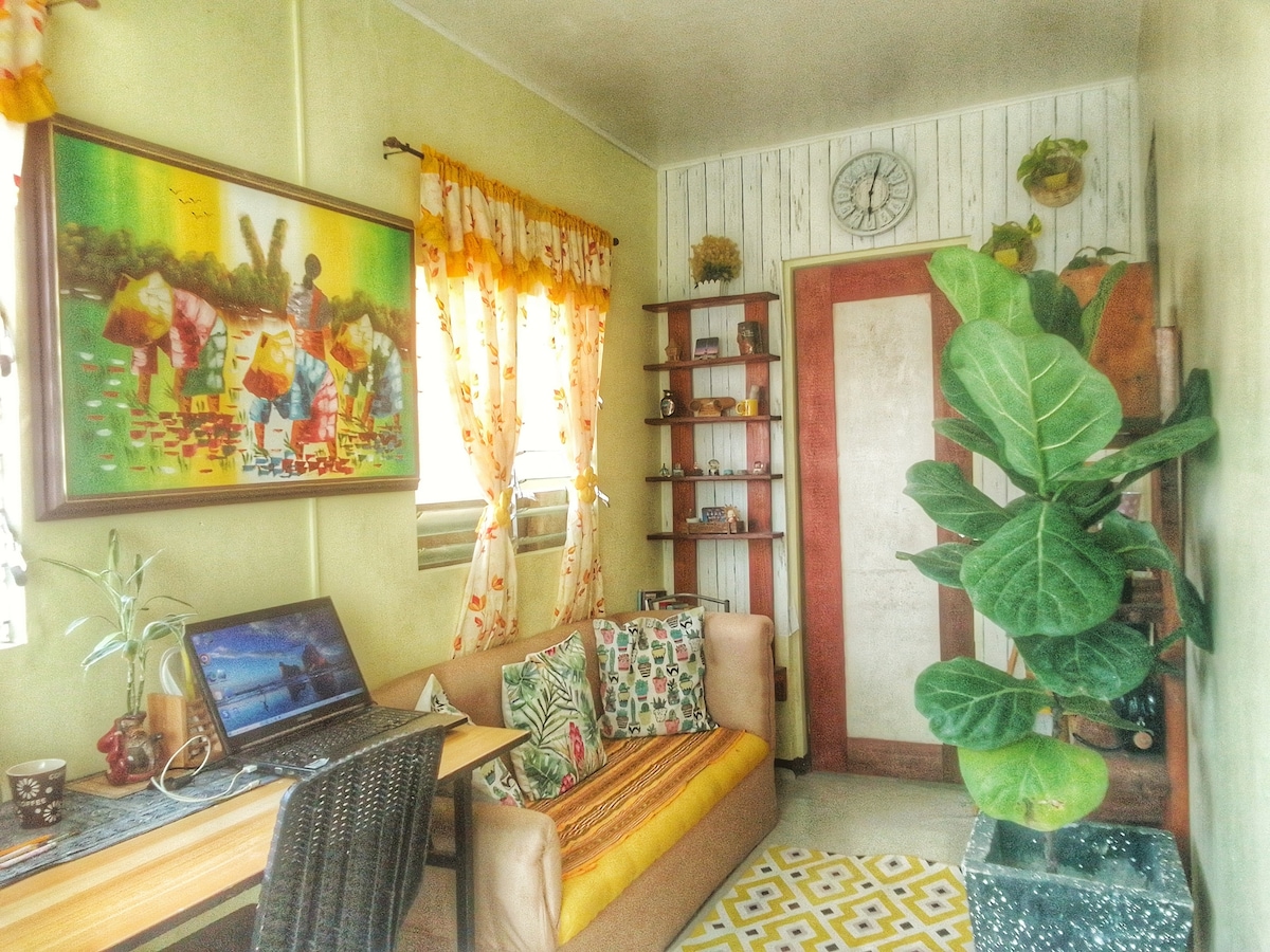A cozy living area is depicted, featuring a combination of a vibrant wall painting and a soft sofa with colorful cushions. A potted plant adds greenery, while a wooden shelf displays decorative items. Natural light enters through window treatments, creating a bright atmosphere.