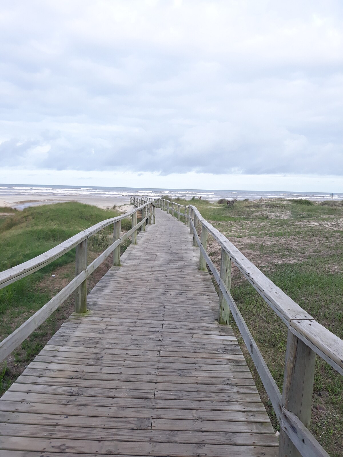 A wooden boardwalk extends toward the beach, surrounded by green grass and coastal vegetation. The pathway leads through a natural landscape, with the ocean visible in the distance beneath a cloudy sky.