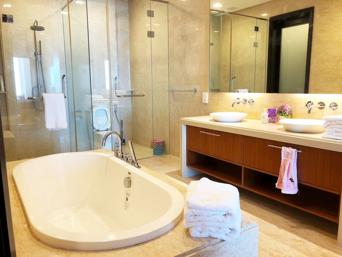A spacious bathroom is featured with a large freestanding tub and a separate glass walk-in shower. Dual sinks are positioned on a wooden vanity, with plush towels neatly arranged nearby. Natural light filters in, enhancing the elegant stone finishes throughout.