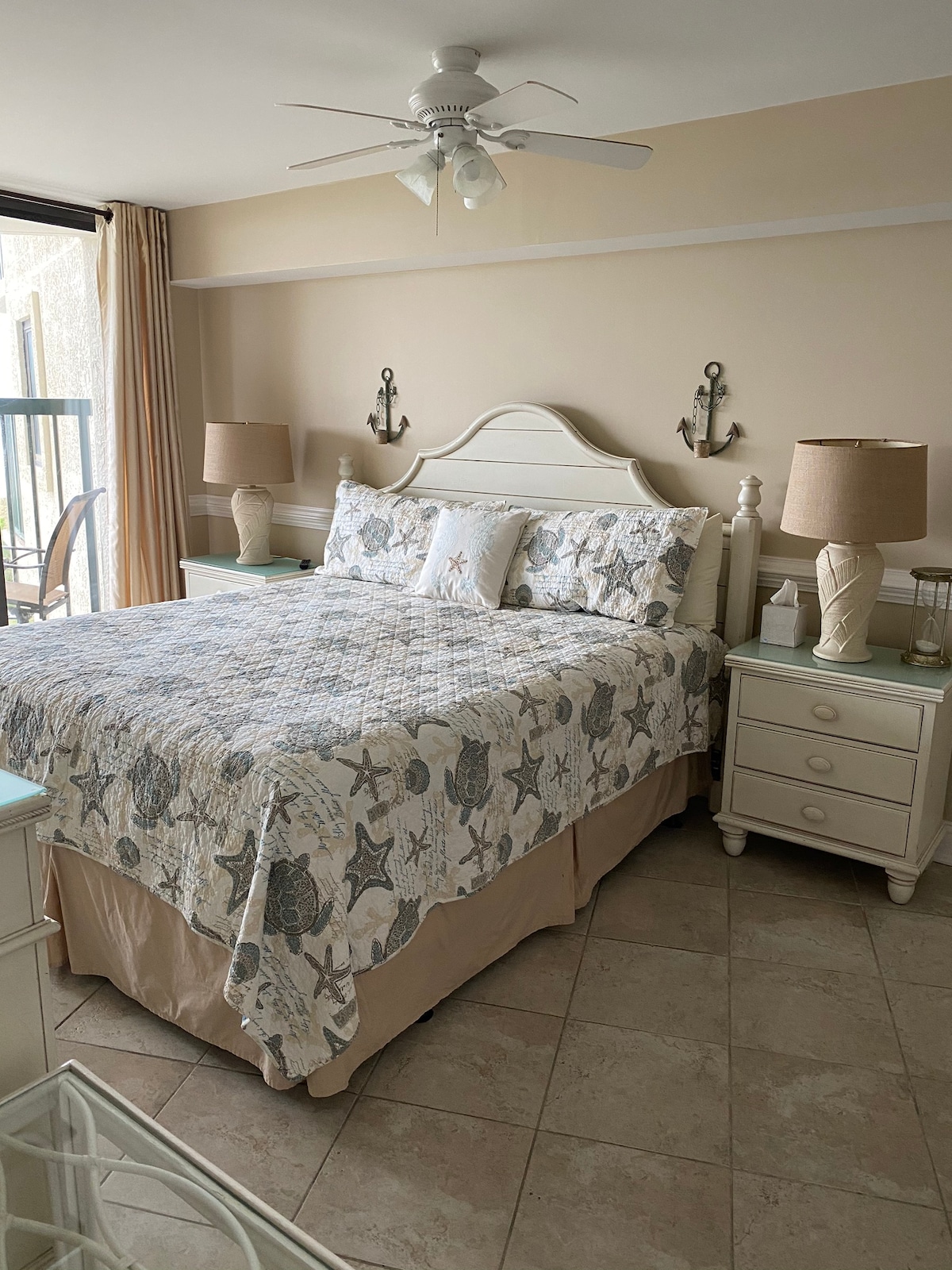 A cozy bedroom is featuring a spacious bed with a starfish-patterned quilt. Two bedside tables with lamps are positioned on either side. Gentle light streams in through a nearby window with sheer curtains, complementing the soft, neutral tones of the decor.