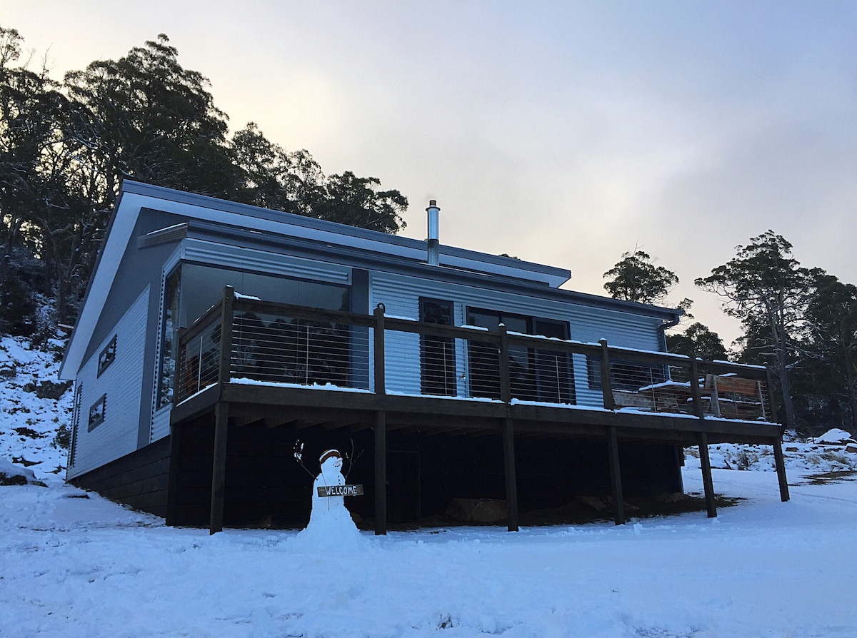 A modern retreat is pictured, featuring a spacious wooden deck surrounded by snow. The exterior is clad in light-colored materials, with large windows that allow natural light. A snowman with a welcoming sign stands in the foreground, adding a cheerful touch to the winter landscape.