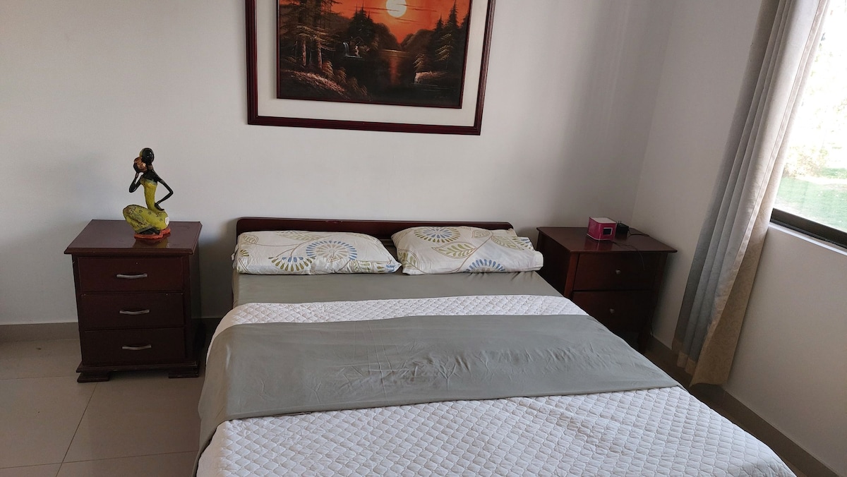 A spacious bedroom is featured with a neatly made bed layered in a light quilt. Nightstands sit on either side, complemented by decorative items. A window provides natural light, enhancing the serene atmosphere created by the framed landscape art on the wall.