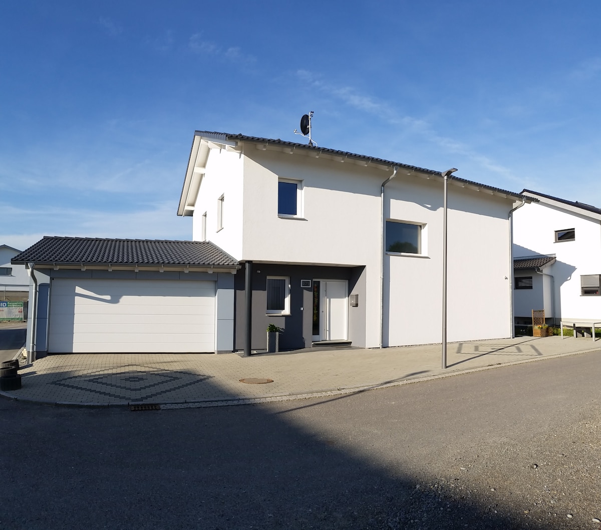 A modern, two-story house is displayed, featuring a clean white facade and a pitched roof. A driveway leads to a spacious garage on the left. A well-maintained entrance area is visible, with a front door framed by minimalistic landscaping. Bright blue skies and a serene setting complete the scene.