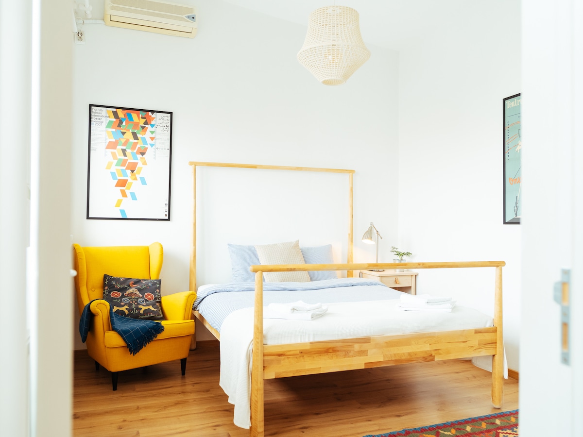 A minimalist bedroom is shown, featuring a spacious double bed with light bedding and two plush towels placed neatly at the foot. A bright yellow armchair adds a pop of color, alongside a small bedside table with a decorative lamp. Soft wooden flooring and white walls create a calming ambiance.