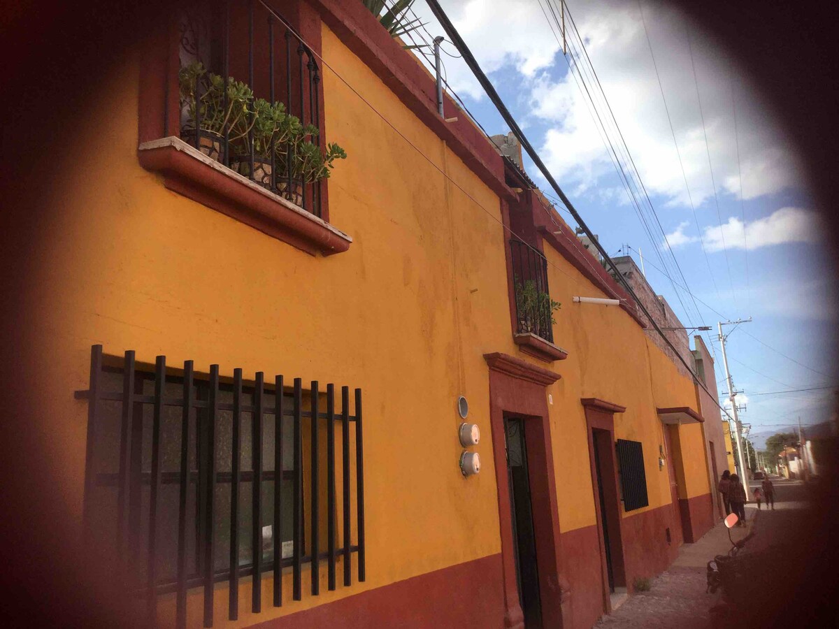 A quaint yellow building with a textured exterior features multiple windows adorned with black iron bars. Small plants can be seen on the balconies. The setting includes a clear blue sky, and the surrounding area hints at a charming neighborhood.