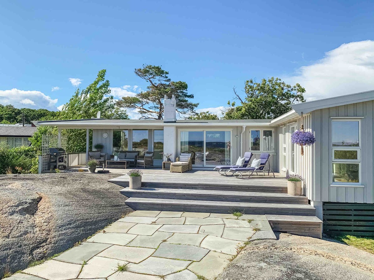 A wooden deck extends from the main entrance of the cabin, featuring comfortable lounge chairs and planters with purple flowers. The large windows allow natural light into the interior. A rocky landscape and greenery are visible in the background, indicating the cabin's proximity to nature.
