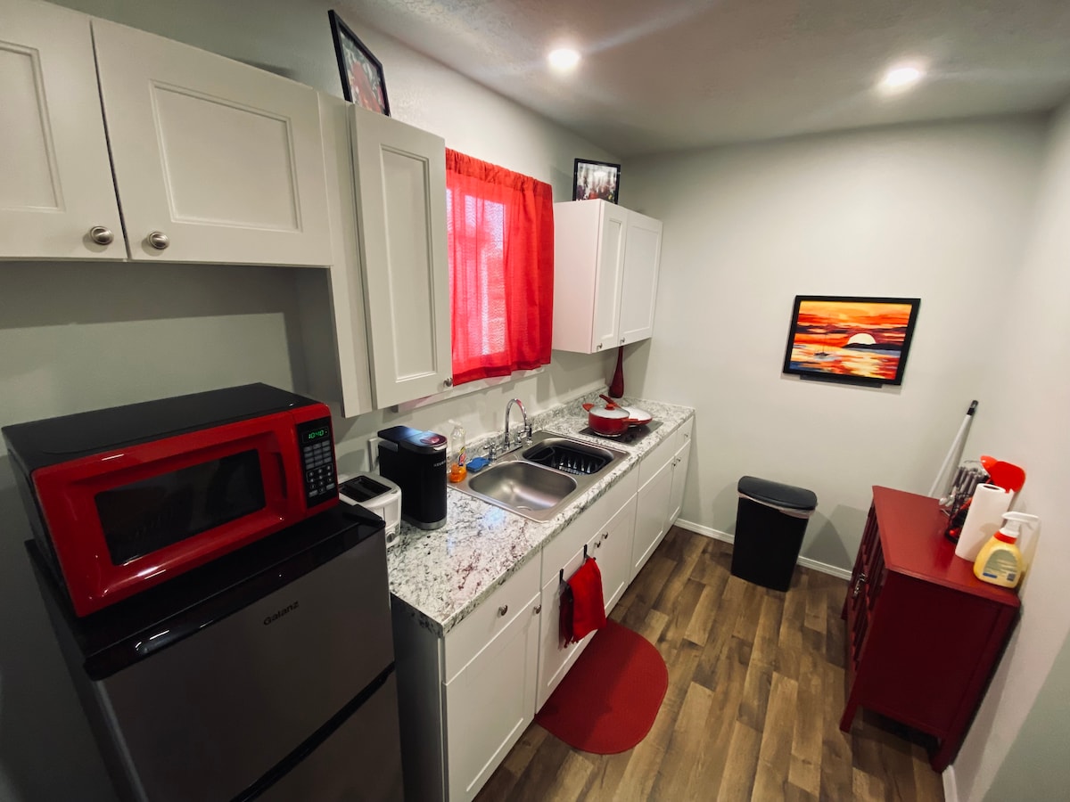 A compact kitchen space is presented with modern white cabinetry, offering a granite countertop with a stainless steel sink. Appliances include a microwave and a bright red microwave, with a black trash can nearby. Light filters in through a window adorned with red curtains.
