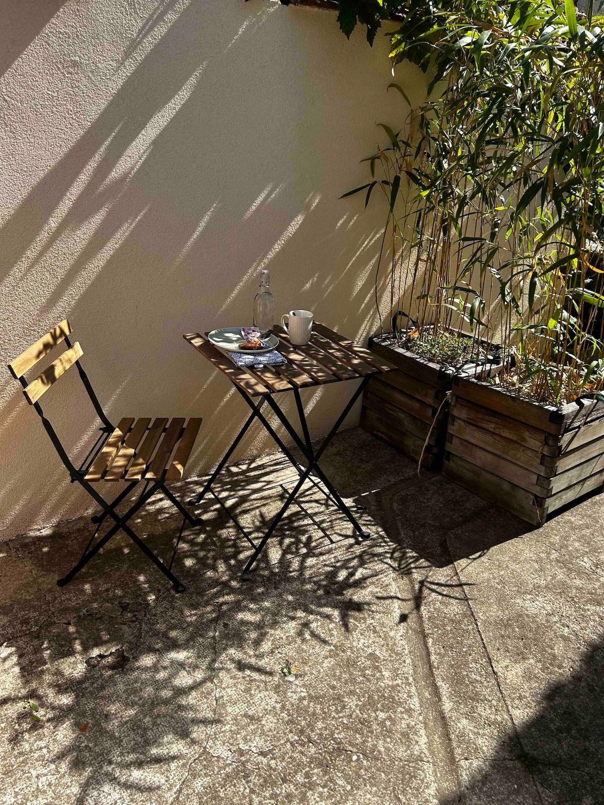 A small outdoor space is depicted with sunlight casting shadows from a nearby wall. A foldable wooden chair accompanies a compact table, which holds a cup and a plate. A wooden planter with green bamboo adds a natural element to the setting.