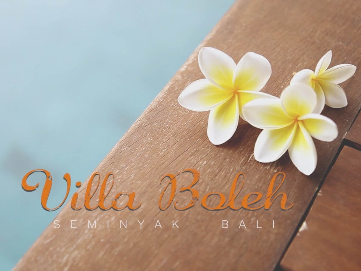 Two delicate white and yellow frangipani flowers are placed on a smooth wooden surface, highlighting the tropical ambiance. The blurred background suggests a serene water feature, evoking a sense of calm associated with the villa's natural surroundings.