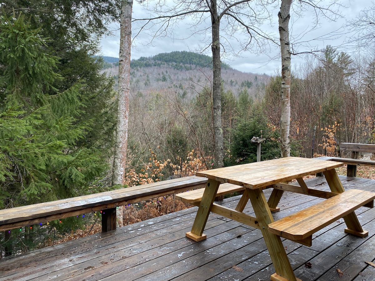 A picnic table is positioned on a wooden deck, surrounded by trees with a view of rolling hills in the background. The deck surface shows a natural wood finish, and the setting appears tranquil, with a few scattered leaves indicating the season.