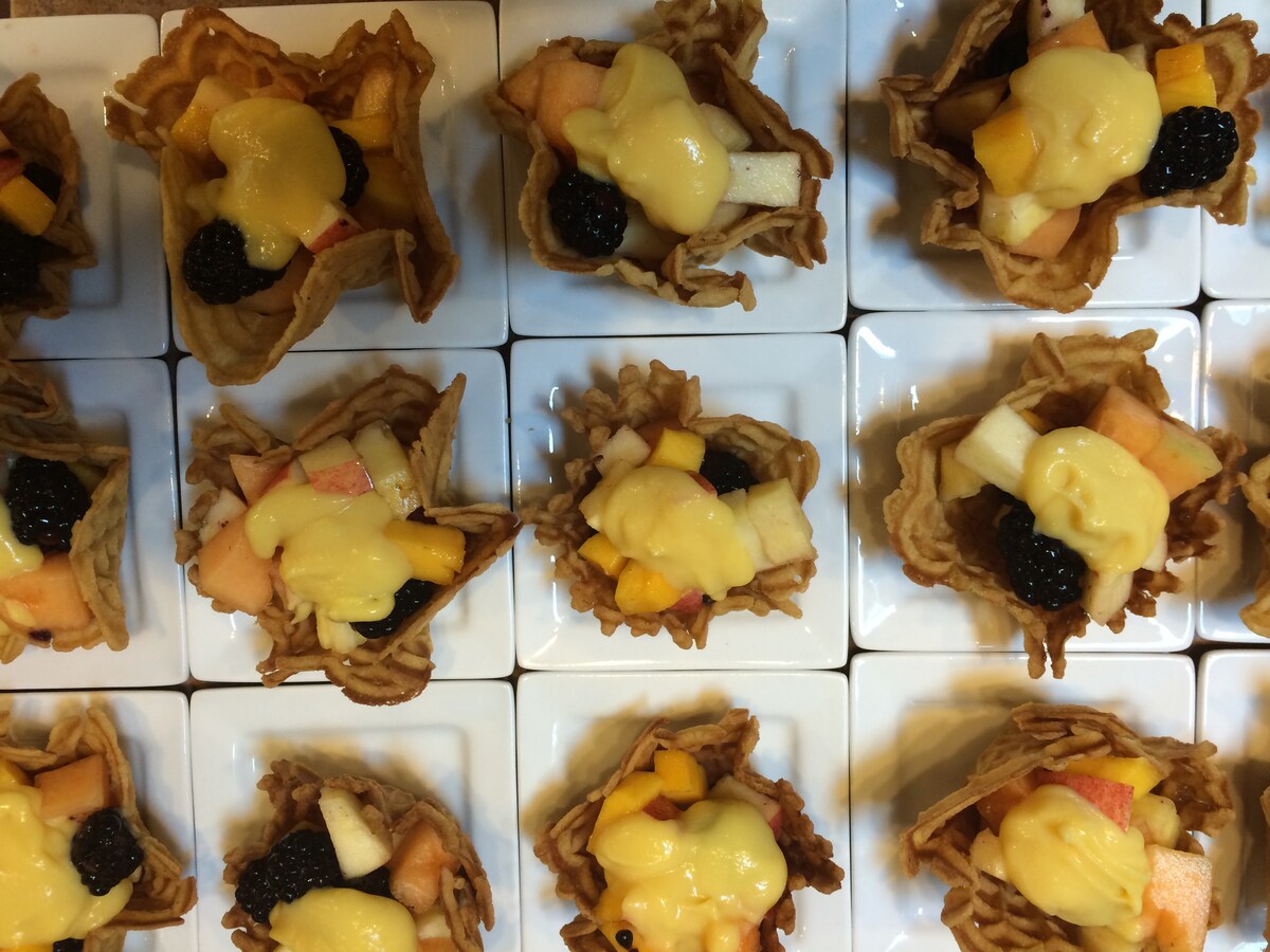 Small, delicate dessert cups are arranged neatly on white plates. Each cup is filled with a variety of fresh fruits, including blackberries and diced apples, topped with a creamy yellow sauce. The presentation highlights the intricate design of the cups.