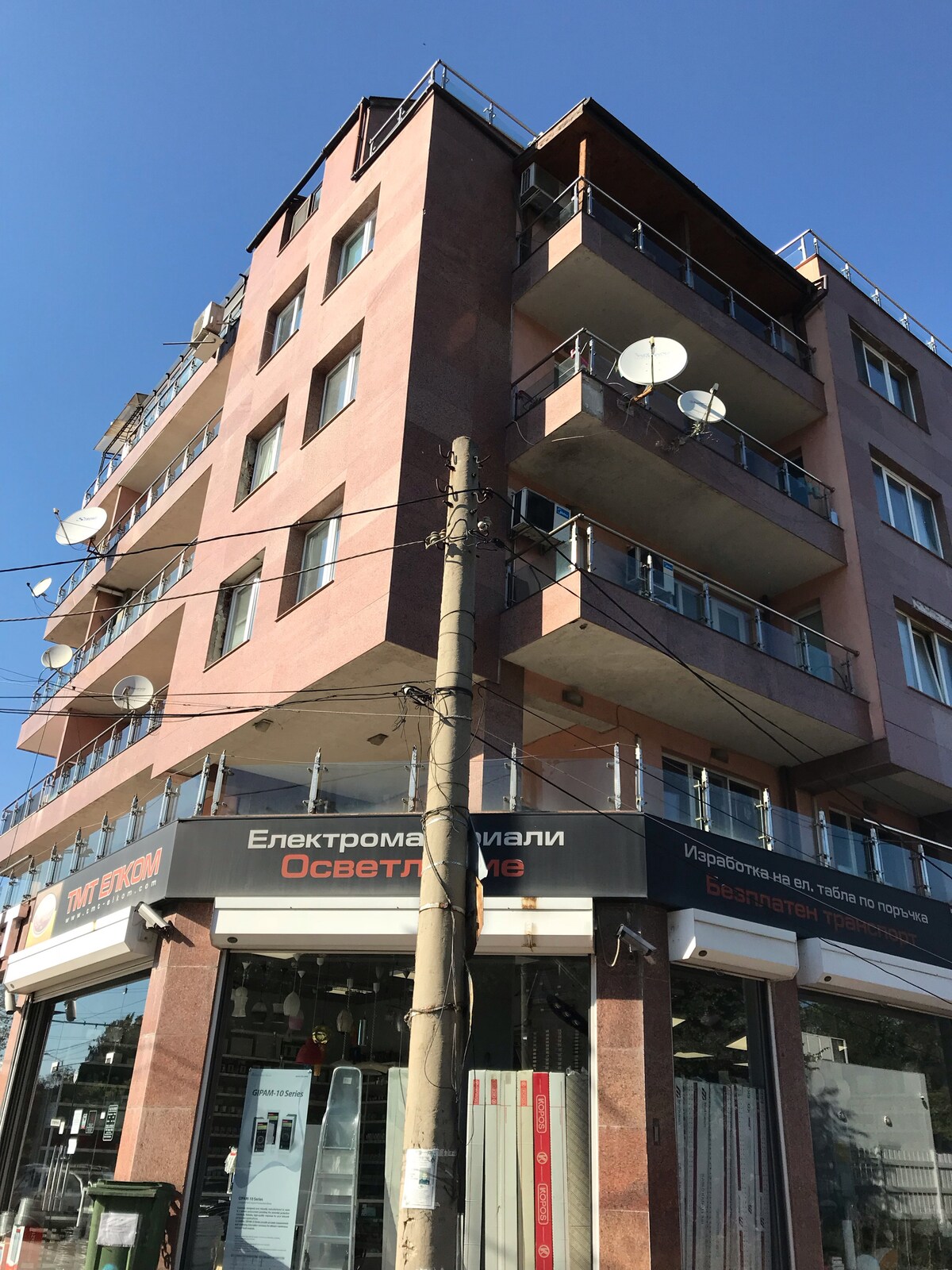 A four-story building showcases a modern façade with large windows and multiple balconies featuring railings. Satellite dishes are mounted on the exterior, while a shop occupies the ground level. Clear skies can be seen in the background, emphasizing the building's urban setting.