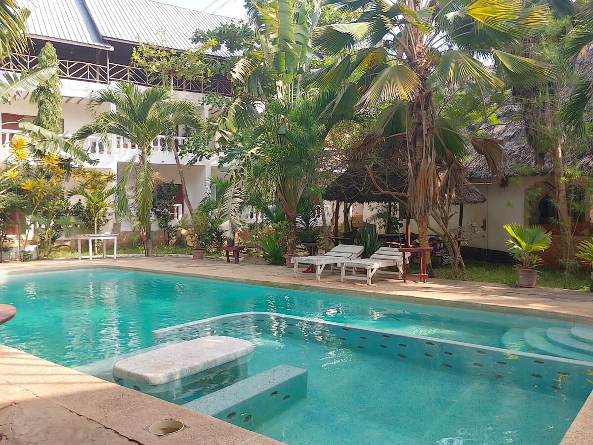 A serene outdoor area features a spacious swimming pool surrounded by lush greenery and tropical plants. Lounge chairs are positioned along the poolside, inviting relaxation. Nearby, a shaded seating area with traditional thatched roofs adds to the tranquil atmosphere.