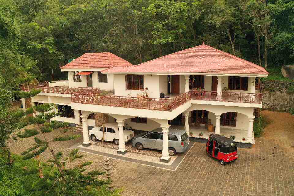 An aerial view of the spacious house surrounded by lush greenery. The structure features a red-tiled roof and multiple balconies, with several vehicles parked beneath. The paved driveway leads to the entrance, creating a welcoming atmosphere.