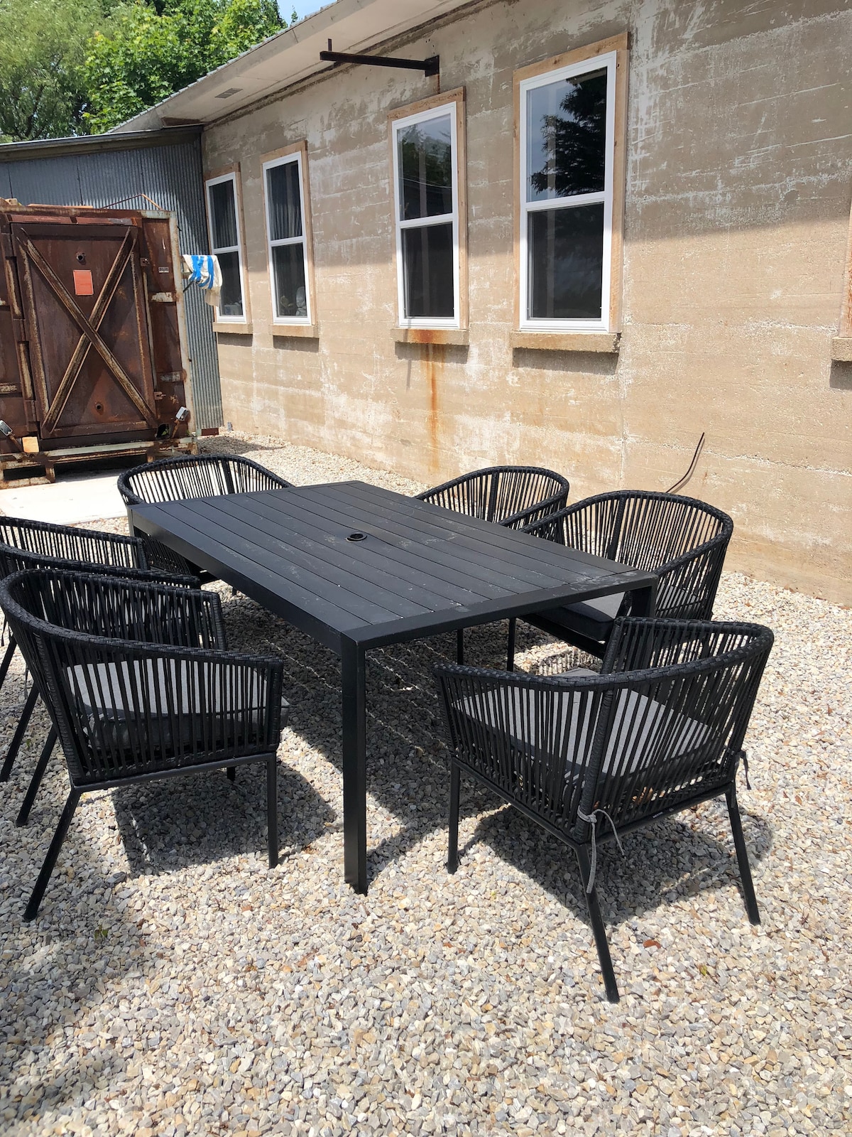 An outdoor dining area is featured, showcasing a rectangular black metal table surrounded by six black wicker chairs with cushioned seats. The setting is placed on a gravel surface next to a textured beige wall, with natural light illuminating the scene.