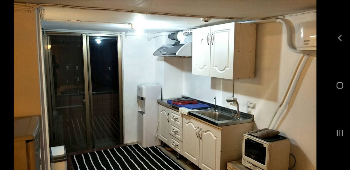 A compact kitchen features white cabinetry and a stainless steel sink, accompanied by essential appliances, including a refrigerator and a small stove. A dark striped rug covers the floor, while natural light enters through sliding glass doors that lead to a balcony.