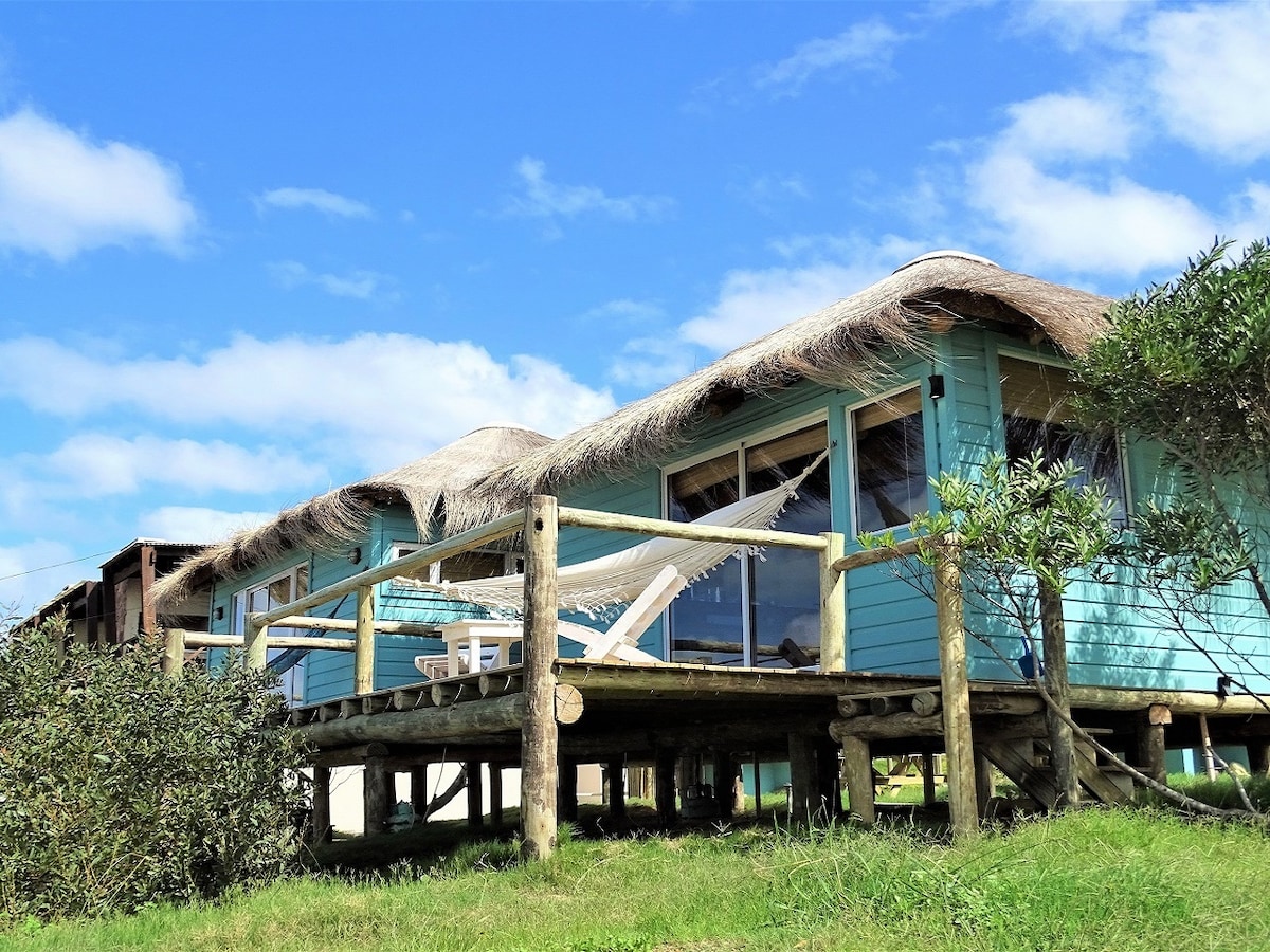 The cabin features a thatched roof and is elevated on wooden stilts. A deck extends from the structure, furnished with a hammock and a small table with chairs. Lush green grass surrounds the property, set against a backdrop of blue sky and fluffy clouds.