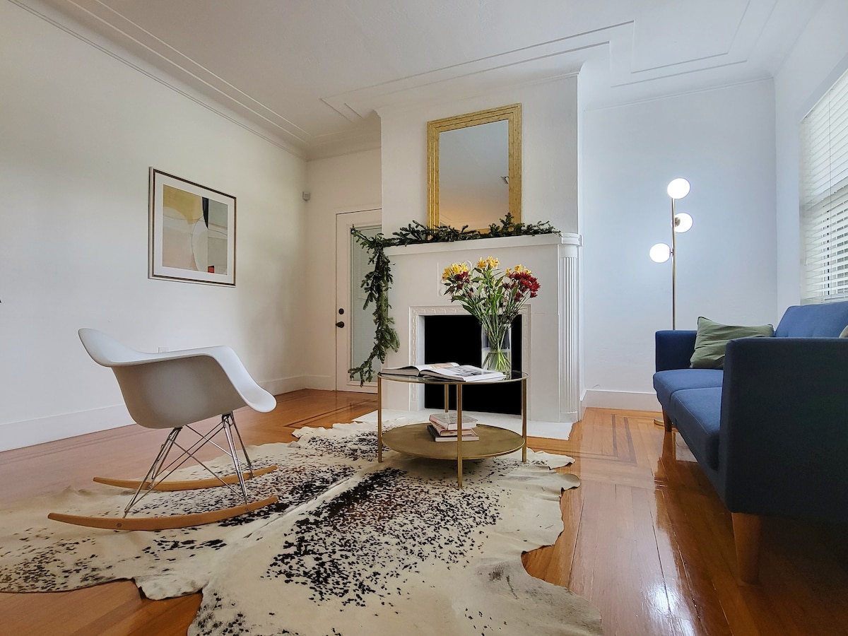 A sunlit living room features a stylish rocking chair and a modern blue sofa. A round coffee table sits atop a cowhide rug, complemented by fresh flowers in a vase. Decorative elements include a framed artwork and a minimalist floor lamp near the fireplace.