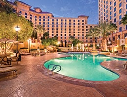 An inviting outdoor pool area is surrounded by lush palm trees and private cabanas. Lounge chairs are neatly arranged along the pool's edge, reflecting a tranquil ambiance, while soft lighting illuminates the space as twilight descends.