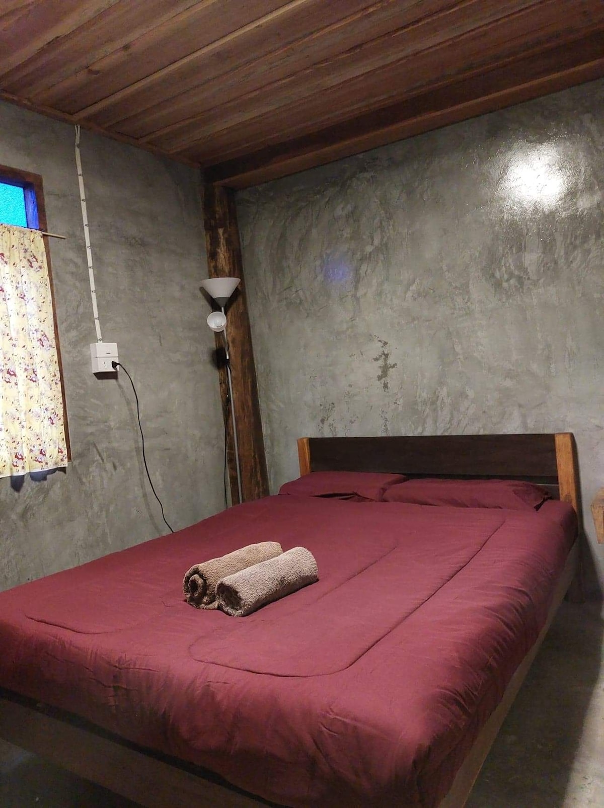 A spacious bed with maroon bedding is centered in a room with exposed wooden beams and a concrete wall. Two rolled towels are placed neatly on the bed. A small window features a floral curtain, allowing natural light to enter the room.