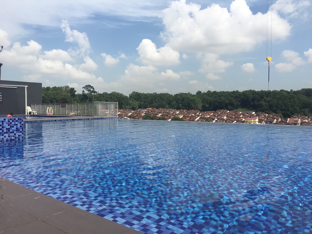 An infinity pool extends towards the horizon, featuring shimmering blue tiles that reflect the sky above. Beneath a partly cloudy sky, a lush green landscape is visible in the background, providing a serene setting for relaxation.