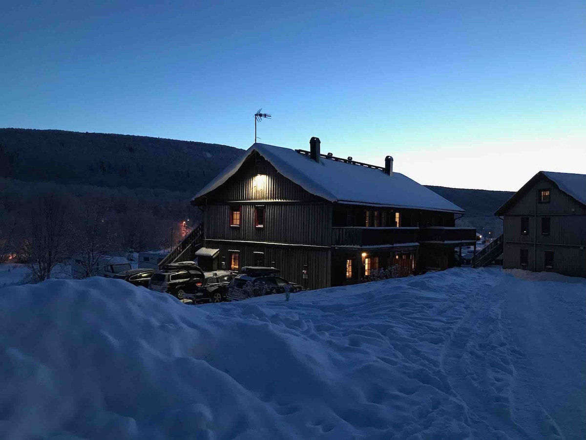 The exterior of the two-story building is shown, surrounded by snow with a clear evening sky. Warm light can be seen emanating from windows, enhancing the welcoming appearance. Nearby vehicles are parked, indicating accessibility. The landscape features snow-covered hills in the background.