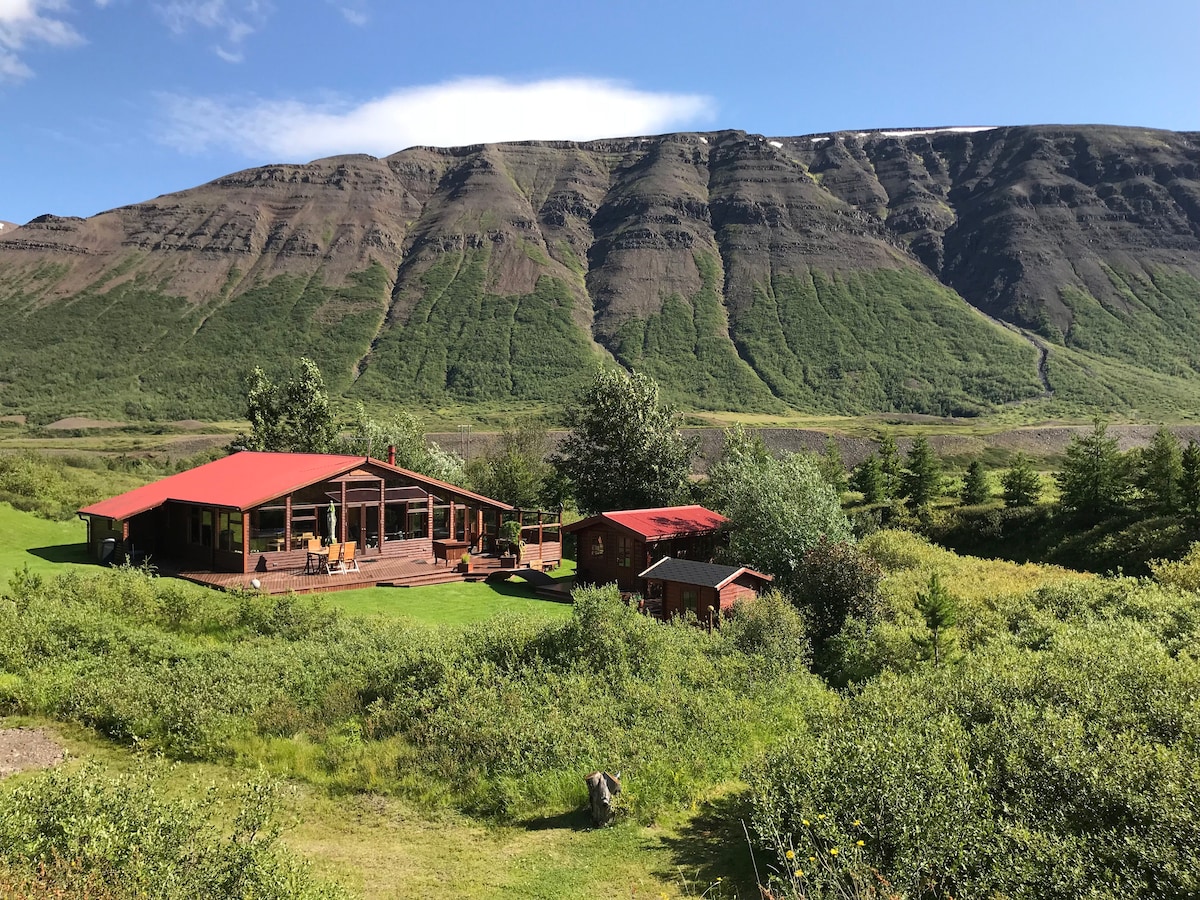 The summer house and guesthouse are set against a backdrop of rugged green hills. The main house features a red roof and large deck, while the surrounding lawn is filled with trees and shrubs, creating a peaceful outdoor space.
